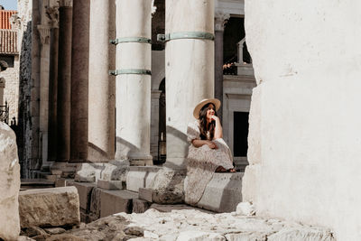 Young woman wearing long dress sitting on stone of ancient ruins in split, croatia.