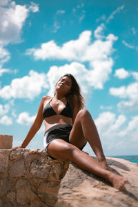 Portrait of woman in bikini sitting on rock against sky