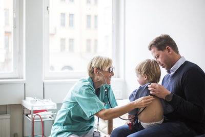 Smiling female doctor examining boy's back with stethoscope while sitting by father in hospital