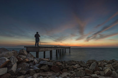 Rear view of man standing on pier over sea against sky during sunset
