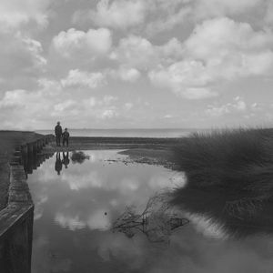 Friends standing by reflection in water at beach against cloudy sky