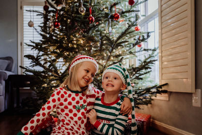 Siblings in costume sitting against christmas tree at home