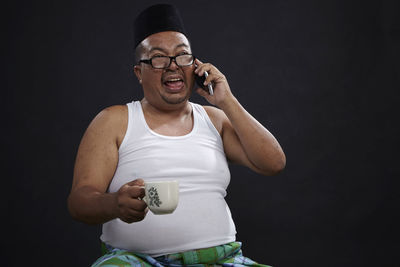 Mature man talking on phone while holding coffee cup against black background