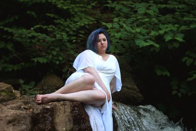 Young woman looking away while sitting on rock by waterfall in forest