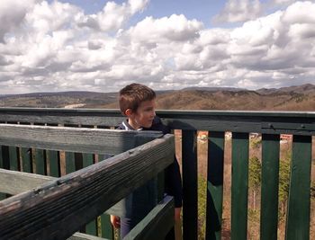 Boy standing on railing against sky