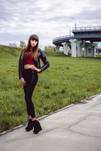 Full length portrait of fashionable young woman standing on footpath by field with bridge in background