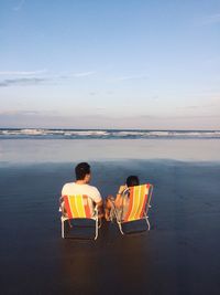 Rear view of men sitting on chair at beach against sky