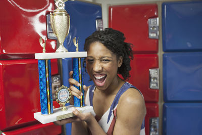A young woman with a trophy in fornt of red and blue lockers.