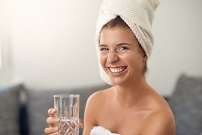 Portrait of smiling young woman wearing bathrobe drinking water while sitting on sofa at home