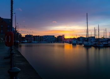 Sailboats moored on harbor by buildings against sky at sunset