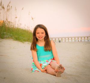 Portrait of smiling girl sitting at beach against sky during sunset