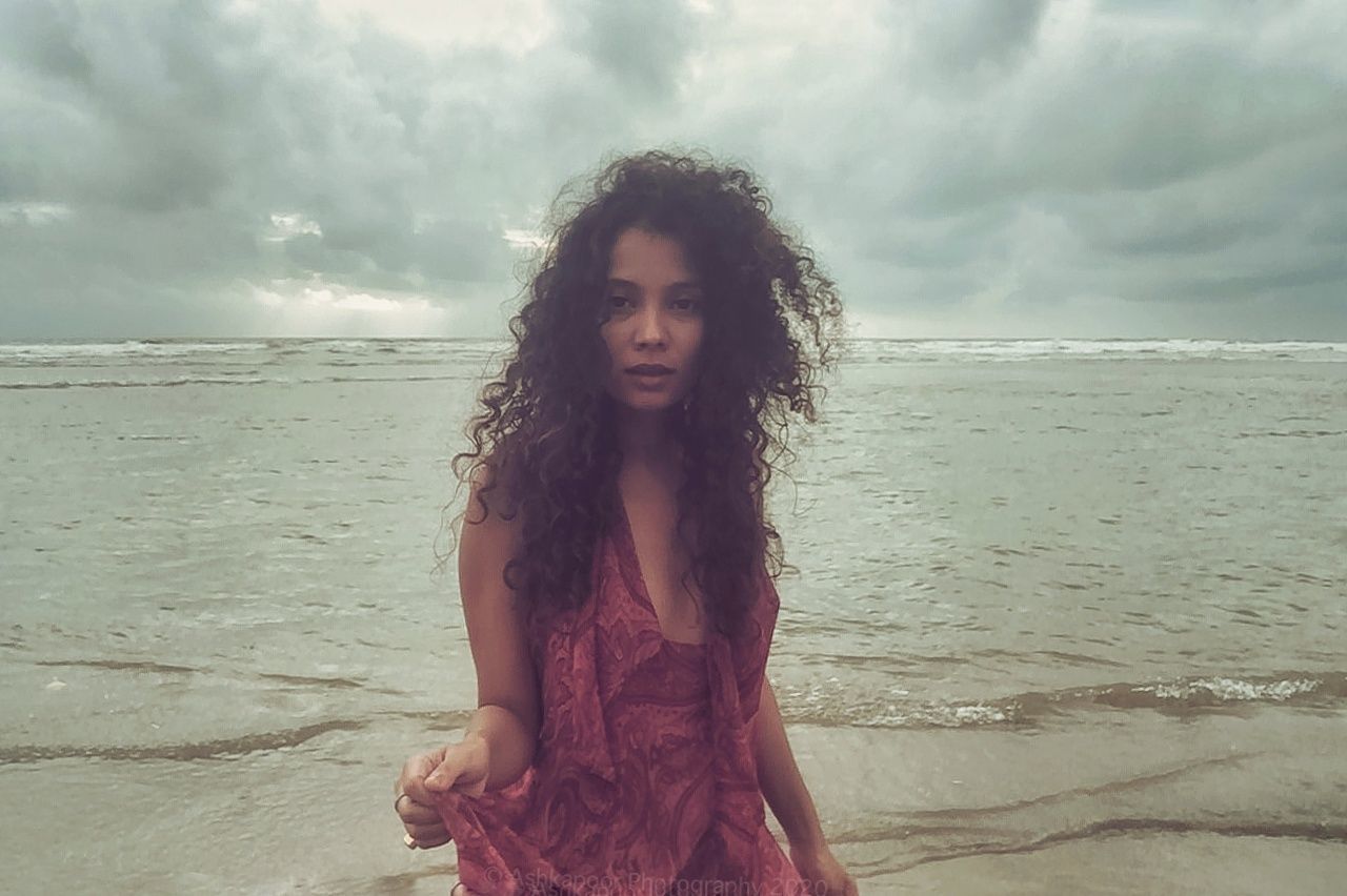 land, hairstyle, long hair, one person, sky, water, sea, curly hair, beach, cloud, nature, young adult, adult, women, brown hair, portrait, beauty in nature, sand, clothing, female, fashion, person, environment, dress, horizon, looking at camera, standing, vacation, holiday, trip, wind, emotion, summer, relaxation, human hair, contemplation, outdoors, leisure activity, horizon over water, front view, looking, smiling, photo shoot, solitude, serious, lifestyles, child, tranquility, overcast, teenager, scenics - nature, wet, happiness