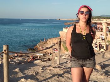 Young woman wearing sunglasses standing at beach against sky