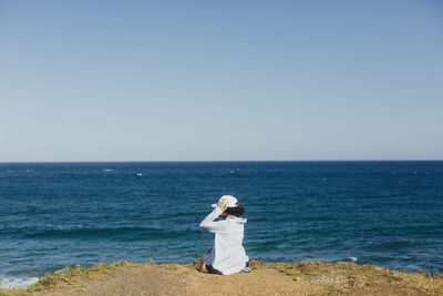 Rear view of woman sitting on cliff by beach against clear sky