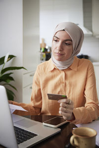 Smiling woman with hijab holding credit card while doing online shopping