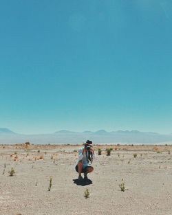 Woman photographing through camera while crouching on field against clear sky