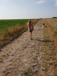 Girl walking with dog on dirt road against sky