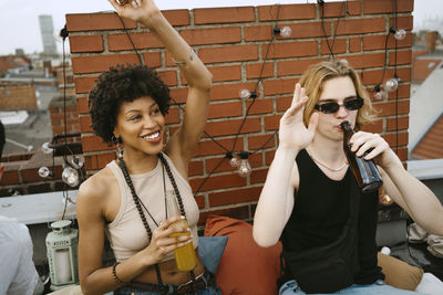 Happy woman and man with hands raised drinking beer on rooftop