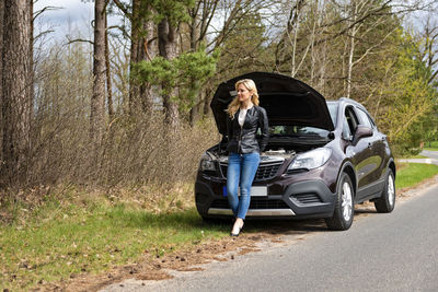 Full length of fashionable young woman standing by breakdown car on roadside