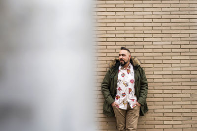 Young bearded man with hands on pocket leaning on wall, looking camera