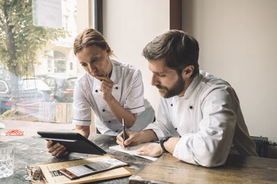 Chef looking at digital tablet while coworker writing at table in restaurant