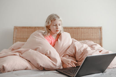 Girl sitting in bed under blanket and learning in virtual online school class. kid working on laptop 