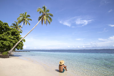 Man with golden hat and green swimsuit at beach with leaning palm tree