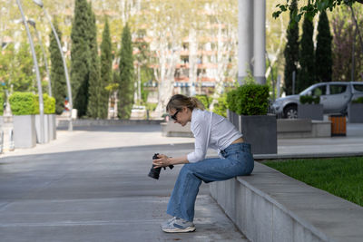 Focused young woman sitting on bench in spring park under tree outdoors resting using camera