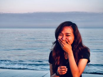 Portrait of beautiful young woman laughing at beach against sky during sunset