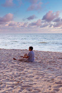 Man sitting on beach by sea against sky during sunset