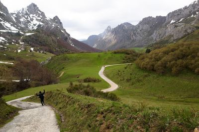 Rear view of person walking on footpath amidst grassy field by mountains
