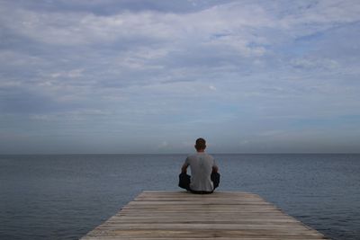 Rear view of man sitting on jetty in sea against sky