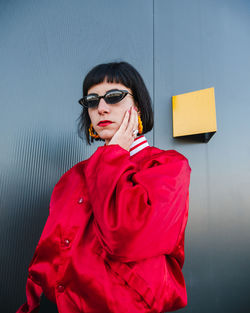 Female millennial wearing vivid red jacket and stylish sunglasses standing with raised arms on street and looking at camera