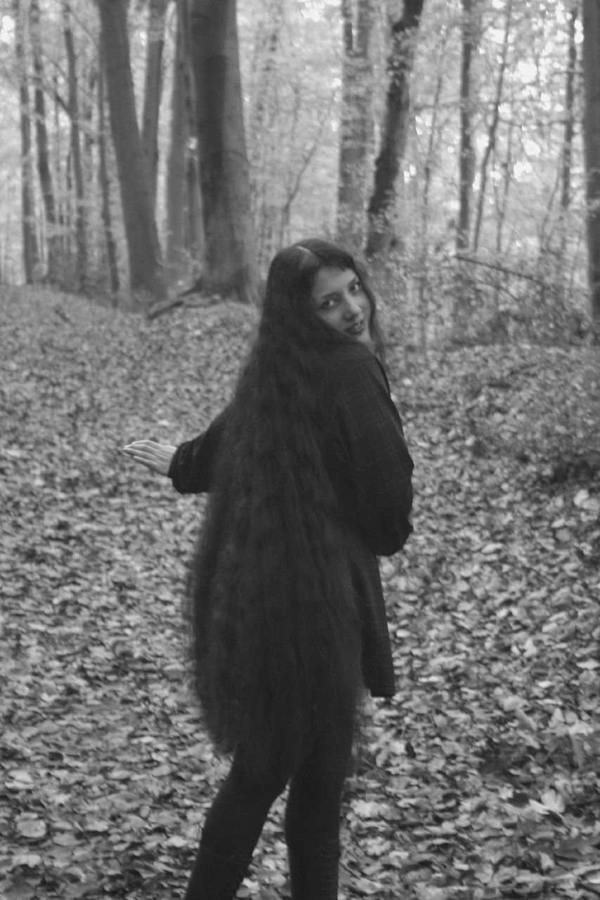 forest, tree, land, black and white, nature, one person, woodland, standing, monochrome photography, plant, tree trunk, one animal, trunk, monochrome, animal, day, black, animal themes, adult, women, clothing, fur, full length, winter, warm clothing, outdoors, young adult, looking, mammal, autumn, portrait, looking away, long hair, non-urban scene