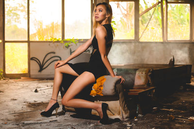 Side view of fashion model sitting on broken armchair in abandoned room