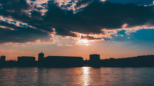 Silhouette buildings by river against sky during sunset