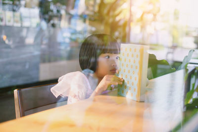 Cute girl reading book on table seen through glass