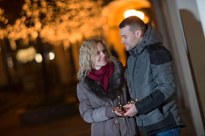 Young couple with illuminated string lights standing in city at night