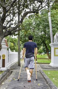 Rear view full length of man walking with crutches on footpath at park
