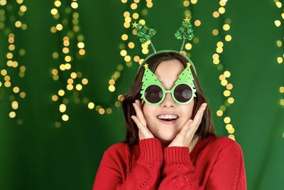 Smiling girl in a red sweater and xmas glasses in the form of christmas trees on a green background
