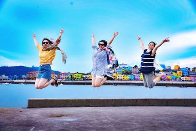 Female friends with arms raised jumping on street amidst sea against blue sky