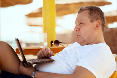 Side view of man with laptop looking away while sitting on chair