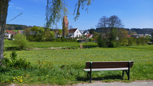 Bench on field by trees and houses against sky
