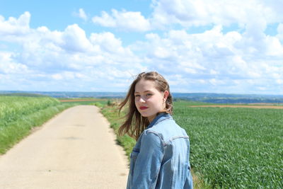 Portrait of woman standing on dirt road amidst field against cloudy sky