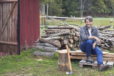 Portrait of man sitting on wooden planks against stacked firewood at yard