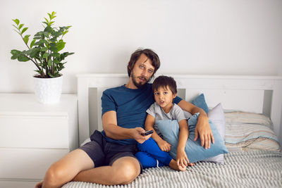 Bearded father and son are sitting on the bed in t-shirts and watching a football match on tv