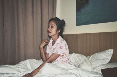 Young woman looking away while sitting on bed at home
