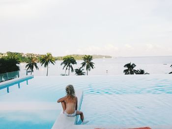 Rear view of girl sitting by swimming pool against sky