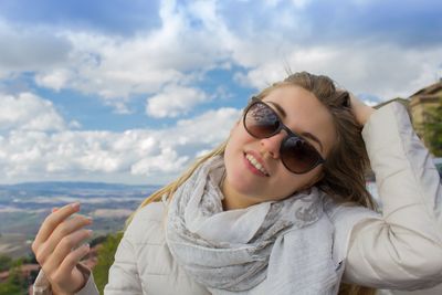 Portrait of smiling young woman with hand in hair against cloudy sky