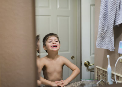 Shirtless smiling boy with broken teeth looking in mirror while standing at home